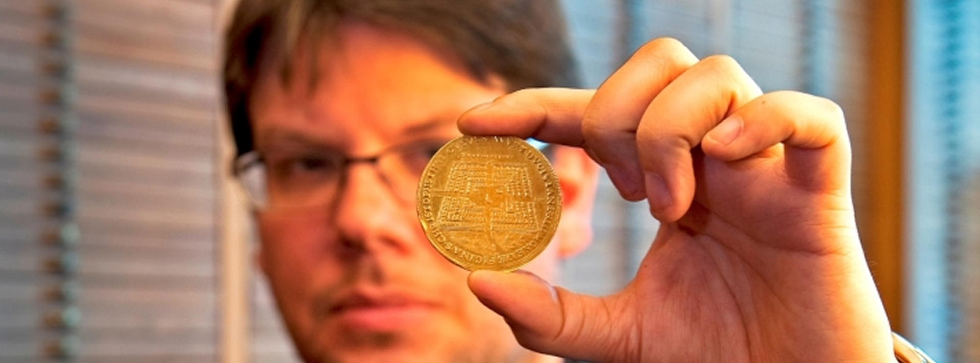 Eine Person mit Brille hält eine große Goldmünze zwischen ihren Fingern und zeigt mit der Vorderseite in Richtung Kamera. Der Hintergrund ist unscharf mit horizontalen Linien.