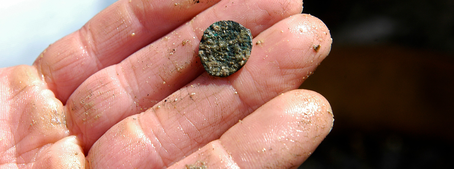 Eine Nahaufnahme einer Person mit sandigen, nassen Fingern, die ein kleines, rundes, korrodiertes Objekt hält, das einer alten Münze oder einem Artefakt ähnelt.